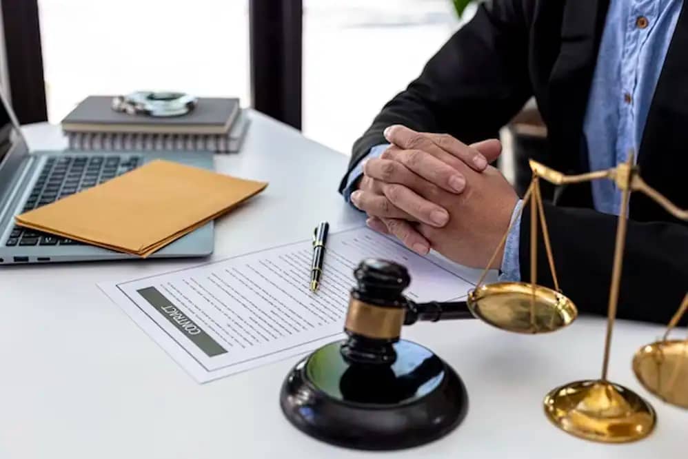 Personal injury lawyer sitting at desk with hands clasped over paperwork with a gavel to his right and a set of justice scales on left