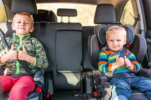 Two little boys sitting on a car seat and a booster seat buckled up in the car.