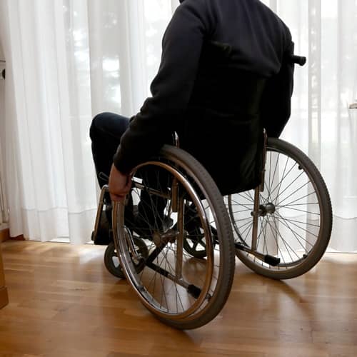 Man in a wheel chair looking out a window through white drapes - Texas Nursing Home Abuse Lawyers