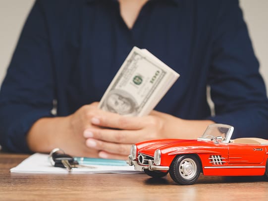 Mujer sosteniendo billetes de $100 mientras estaba sentada en un escritorio con un Corvette de juguete rojo - Subrogación de seguros de automóviles en Texas en reclamaciones por daños a la propiedad
