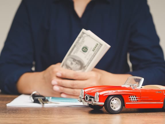 Woman holding $100 bills while seated at a desk with a red toy Corvette