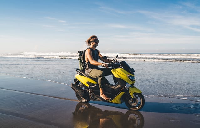 Full length of male traveler in glasses riding motorcycle on wet sand near waving sea while looking away and resting during vacation.