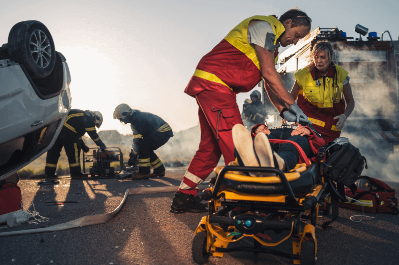 Medic doing cardiopulmonary resuscitation on a person laying down prone on a gurney while fire fighters try to extinguish a fire from a rollover auto accident. Pain and suffering in personal injury cases.