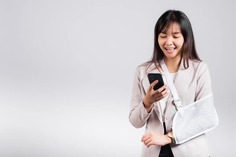 Woman confident smiling broken arm after accident and wear arm splint for treatment and hold smartphone, happy Asian female sling supported hand isolated on white background, social media, copy space. Social Media and Personal Injury Claims