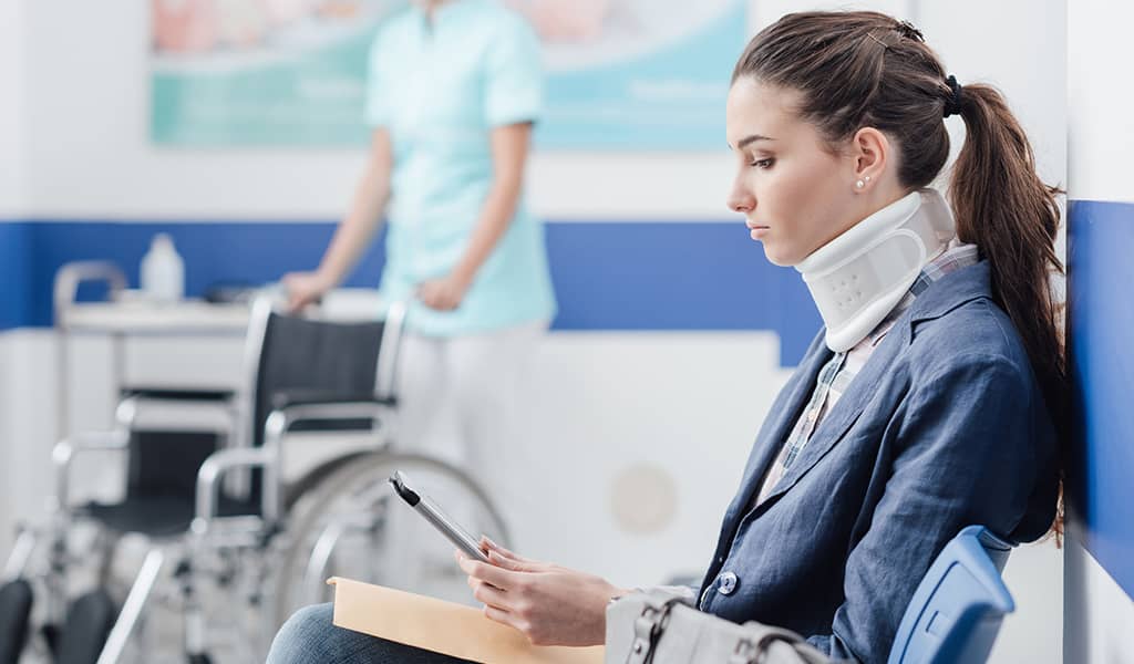 Young female patient with cervical collar support at the hospital, she is sitting in the waiting room and connecting with a digital tablet, medical staff working on the background. Personal Injury Attorney