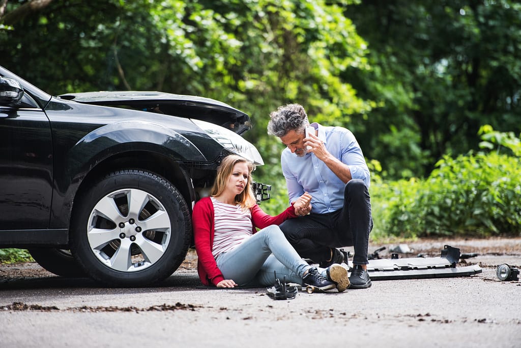 Young woman by the car after an accident and a man with smartphone, making a phone call. Injury