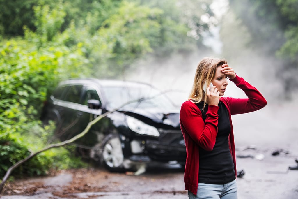 A frustrated young woman with smartphone by the damaged car after a car accident, making a phone call. Claim
